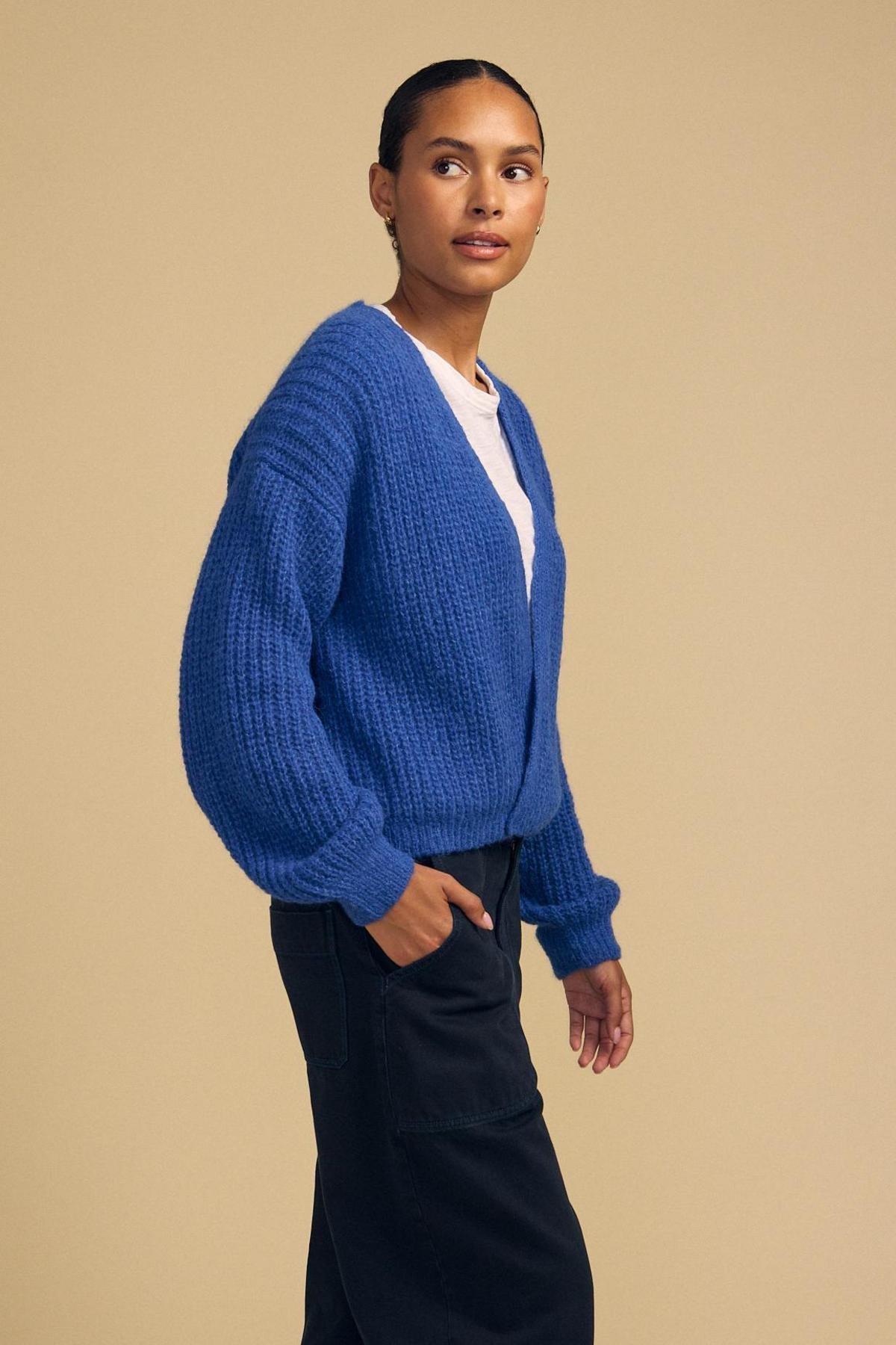 A person stands sideways against a plain beige background, embracing cozy layering in the RANYA ALPACA CARDIGAN by Velvet by Graham & Spencer—a blue alpaca blend cardigan styled over a white top and dark pants.-42181909807297
