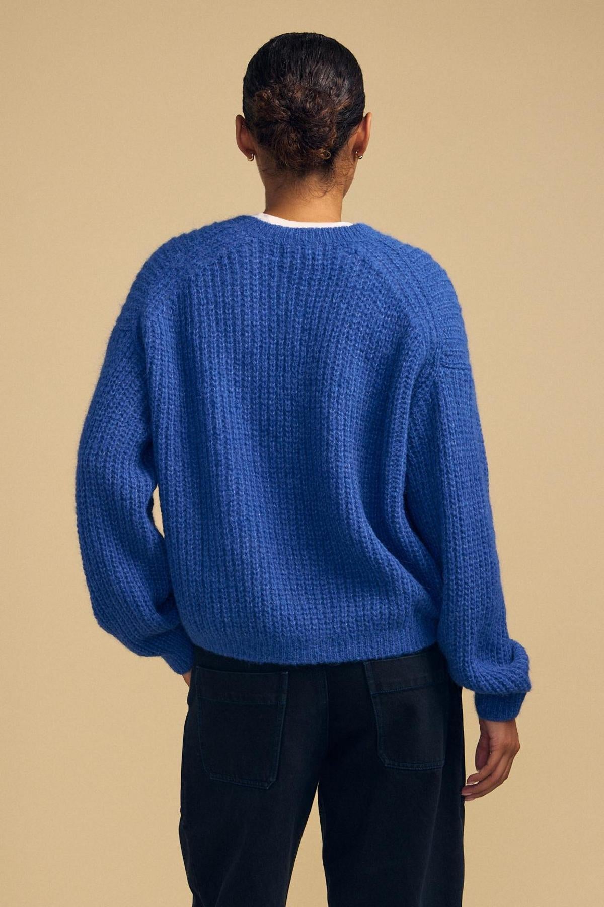 A person stands with their back to the camera against a plain beige background, wearing Velvet by Graham & Spencer’s RANYA ALPACA CARDIGAN—a textured blue alpaca blend layering piece—paired with black pants.