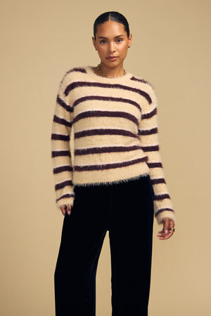 Model wearing a TINA FEATHER YARN SWEATER in a cozy, long-sleeved, fuzzy style with horizontal brown stripes on a cream background and dark trousers.