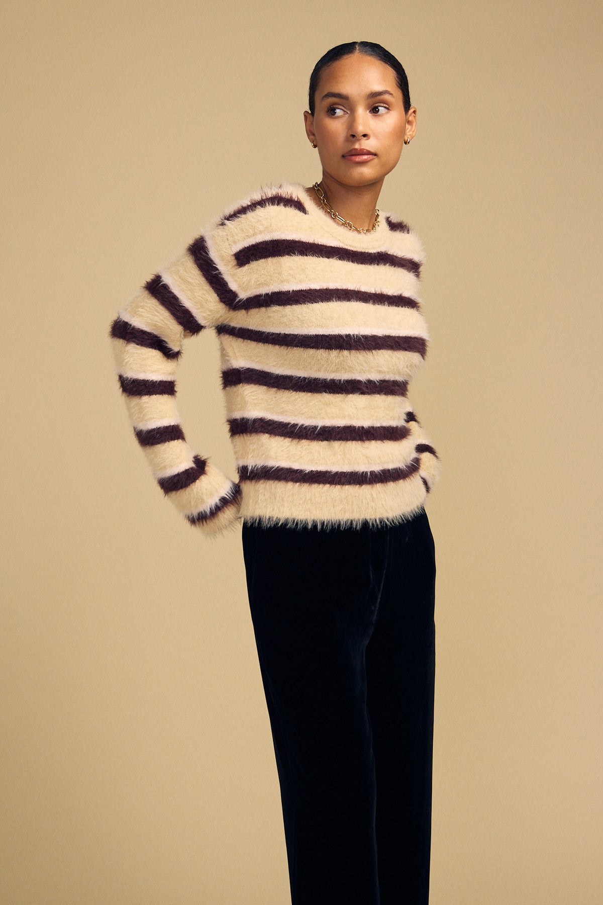 A person wearing a cozy, cream-colored TINA FEATHER YARN SWEATER with horizontal dark brown stripes and dark trousers.
