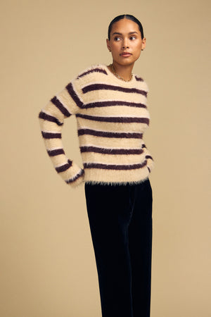 A person wearing a cozy, cream-colored TINA FEATHER YARN SWEATER with horizontal dark brown stripes and dark trousers.