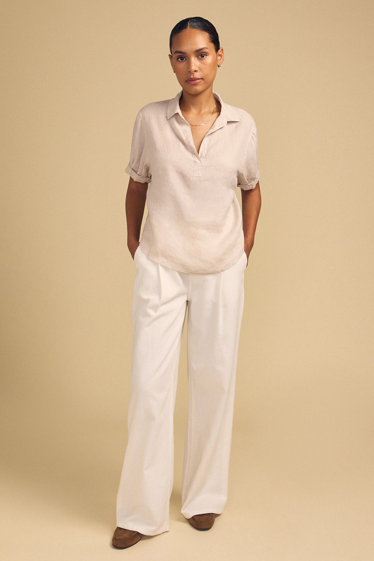 A person stands against a beige background, wearing the VIVICA LINEN TOP by Velvet by Graham & Spencer—a lightweight linen piece featuring a split neckline—paired with white wide-leg pants and brown shoes, hands in their pockets for an effortless look perfect for warm-weather dressing.