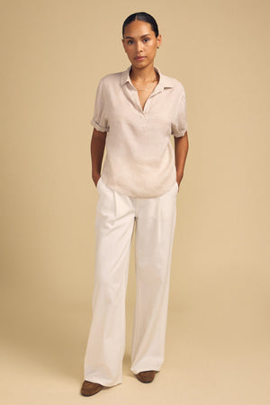 A person stands against a beige background, wearing the VIVICA LINEN TOP by Velvet by Graham & Spencer—a lightweight linen piece featuring a split neckline—paired with white wide-leg pants and brown shoes, hands in their pockets for an effortless look perfect for warm-weather dressing.