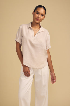 A person stands against a beige background, wearing the VIVICA LINEN TOP by Velvet by Graham & Spencer—a light beige short-sleeve blouse with a split neckline—paired with white pants for an effortlessly chic warm-weather look. One hand rests by their side while the other is slightly raised.
