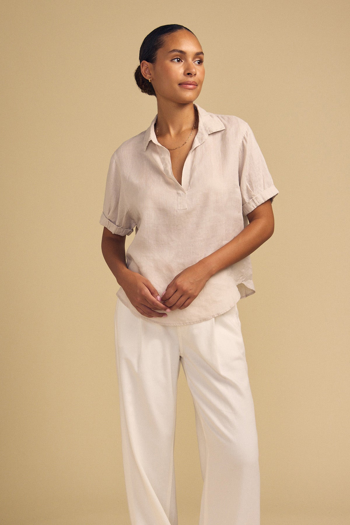 Standing against a beige background, she wears the VIVICA LINEN TOP by Velvet by Graham & Spencer—a lightweight style with a split neckline—paired with white wide-leg pants and her hands loosely clasped, creating a perfect ensemble for warm-weather dressing.-42182020694209