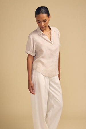 Against a beige background, a woman models the VIVICA LINEN TOP by Velvet by Graham & Spencer—a lightweight linen piece featuring a split neckline—paired with loose white pants, exuding refined ease for effortless warm-weather dressing.