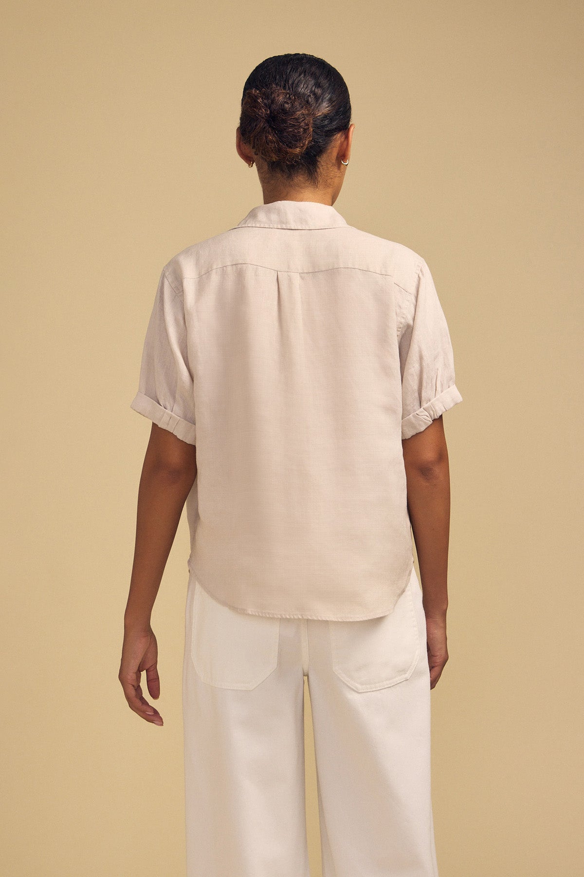 Seen from behind, the person is wearing the VIVICA LINEN TOP by Velvet by Graham & Spencer—its lightweight linen fabric and short sleeves effortlessly paired with white pants against a plain beige background, making it perfect for warm-weather dressing.-42182020628673