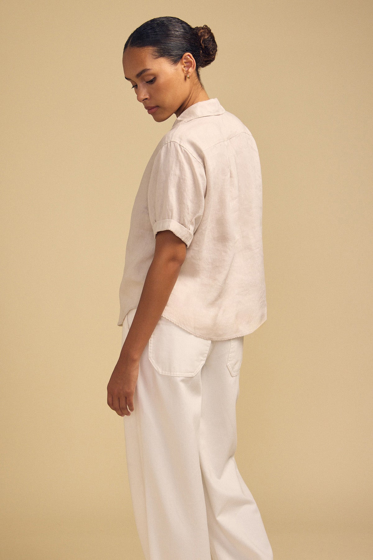 A person stands sideways facing left, wearing the VIVICA LINEN TOP by Velvet by Graham & Spencer—a cream short-sleeve button-up shirt with a split neckline—paired with loose white pants, effortlessly embodying warm-weather style against a plain beige background.