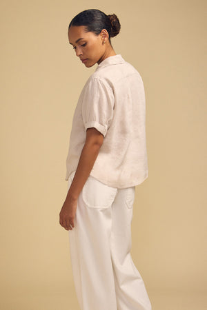 A person stands sideways facing left, wearing the VIVICA LINEN TOP by Velvet by Graham & Spencer—a cream short-sleeve button-up shirt with a split neckline—paired with loose white pants, effortlessly embodying warm-weather style against a plain beige background.