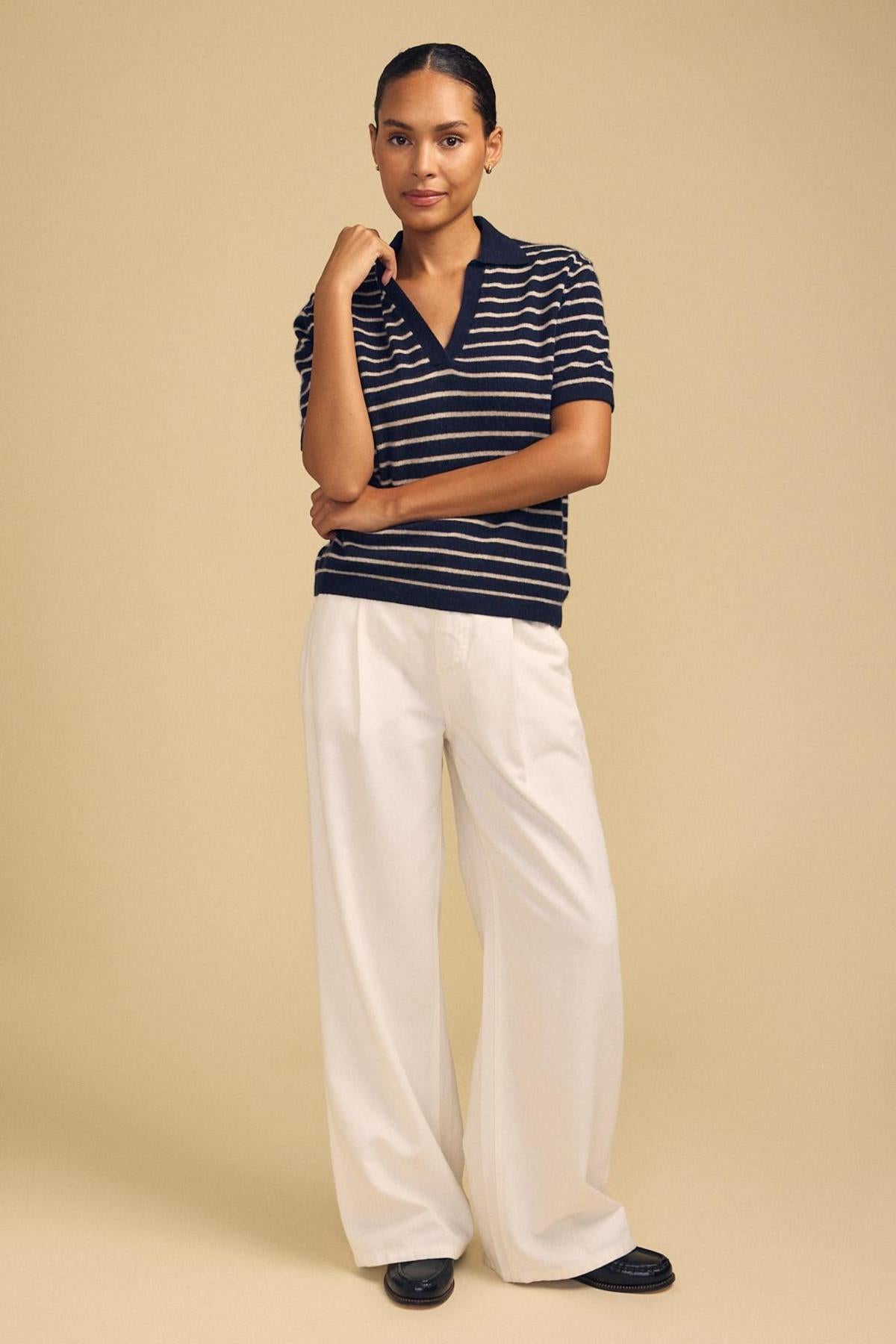 A person stands against a beige background wearing the Velvet by Graham & Spencer YAEL Cashmere Sweater Polo—a navy, striped, short-sleeve, cropped polo—paired with white wide-leg pants and black shoes, one arm bent with hand touching chin.