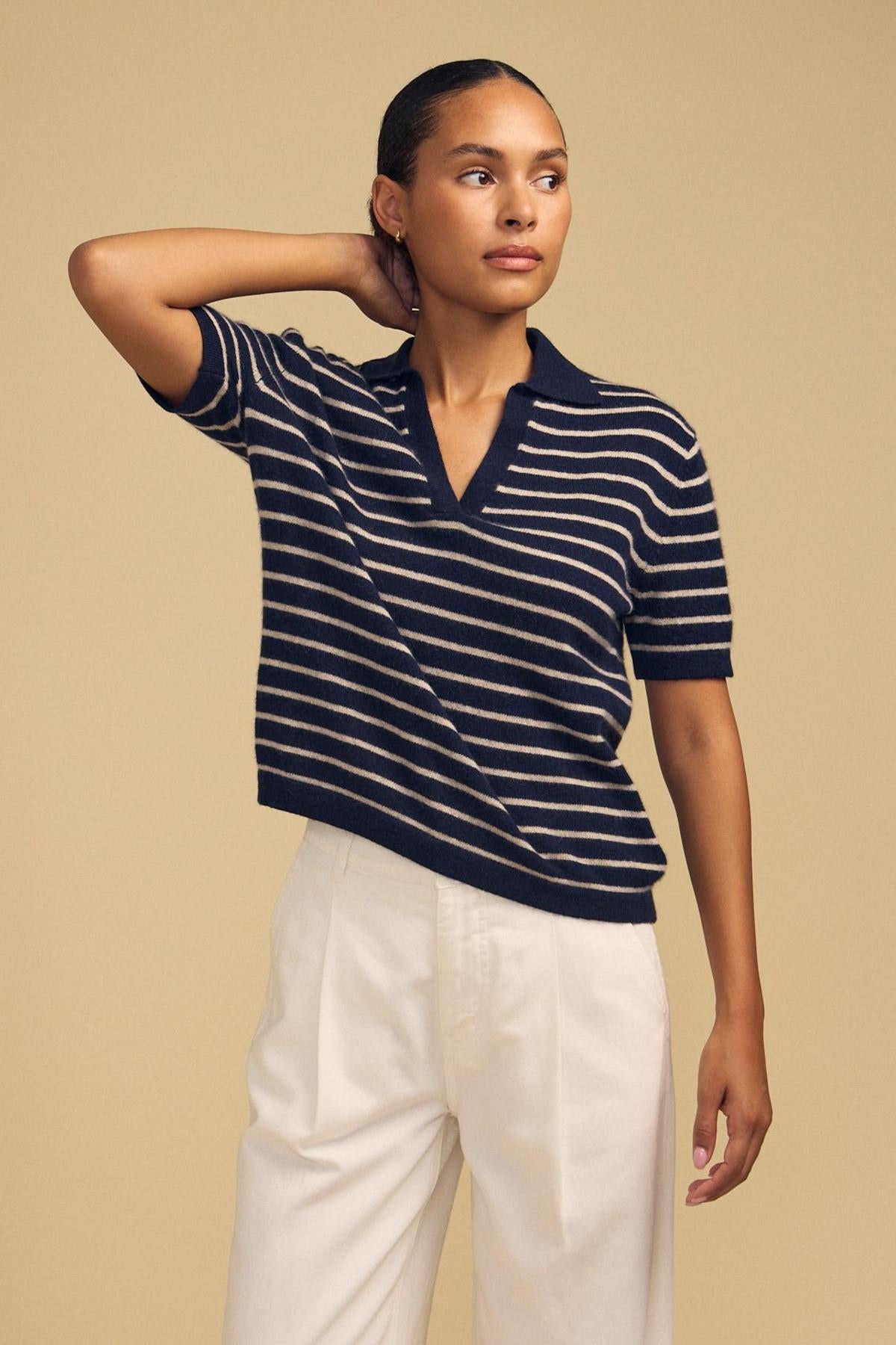 A woman stands against a beige background, wearing the Velvet by Graham & Spencer YAEL Cashmere Sweater Polo in navy blue and white stripes paired with high-waisted white pants, one arm bent with her hand resting behind her neck.