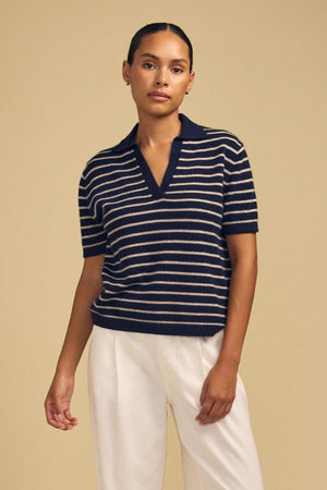 A person stands against a beige background wearing the YAEL CASHMERE SWEATER POLO by Velvet by Graham & Spencer—a navy and white striped cashmere polo with short sleeves and a collar—elegantly paired with white pants.