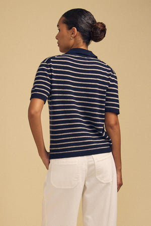 A person stands with their back to the camera wearing the YAEL CASHMERE SWEATER POLO by Velvet by Graham & Spencer, a navy and white striped cropped polo sweater, styled with white pants against a plain beige background.