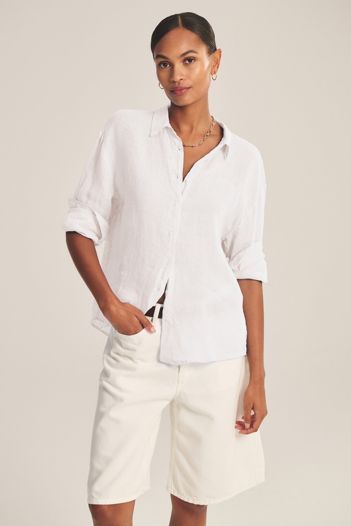 A woman stands against a neutral background wearing the BRIGHTON LINEN SHIRT by Velvet by Graham & Spencer, styled with rolled-up sleeves and paired with knee-length white shorts.