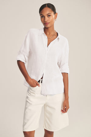A woman stands against a neutral background wearing the BRIGHTON LINEN SHIRT by Velvet by Graham & Spencer, styled with rolled-up sleeves and paired with knee-length white shorts.