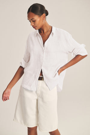 A person wearing the Velvet by Graham & Spencer BRIGHTON LINEN SHIRT, a relaxed fit white button-up with rolled sleeves, paired with knee-length white shorts, stands against a plain background.