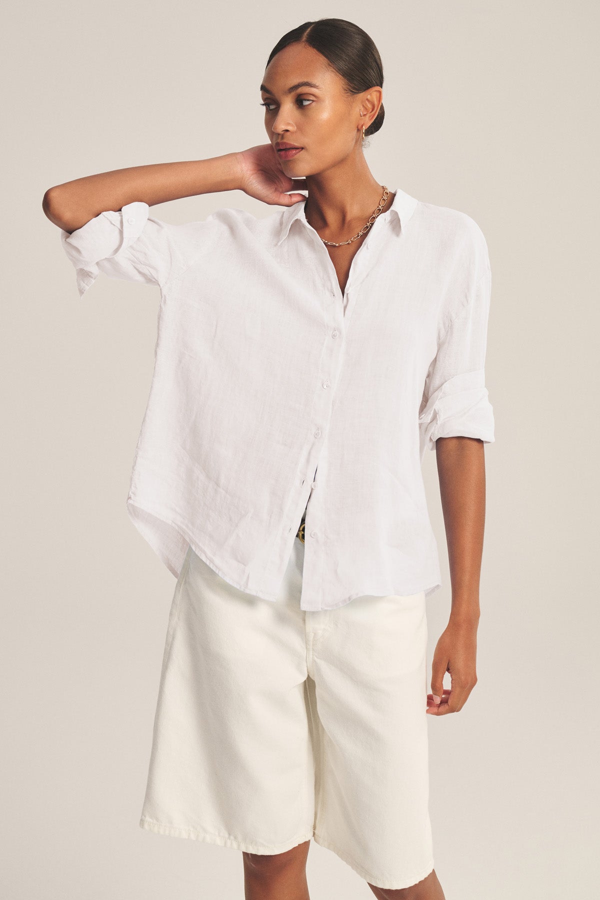 A woman wears the BRIGHTON LINEN SHIRT by Velvet by Graham & Spencer— a relaxed-fit white button-up with rolled sleeves—paired with white knee-length shorts, standing against a plain background.