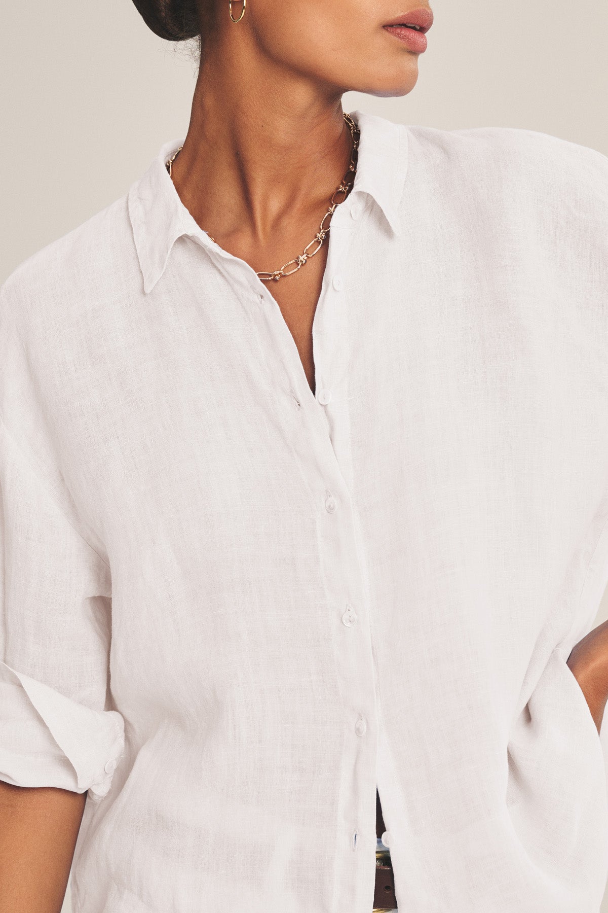A person wearing the BRIGHTON LINEN SHIRT by Velvet by Graham & Spencer—a relaxed fit white woven linen button-up with rolled sleeves—paired with a gold chain necklace and small hoop earring.