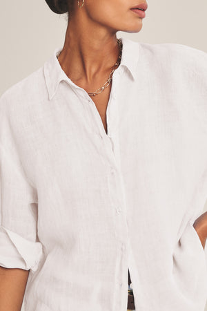 A person wearing the BRIGHTON LINEN SHIRT by Velvet by Graham & Spencer—a relaxed fit white woven linen button-up with rolled sleeves—paired with a gold chain necklace and small hoop earring.