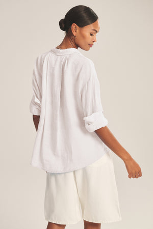Woman wearing the BRIGHTON LINEN SHIRT by Velvet by Graham & Spencer—a relaxed fit, loose white button-up with rolled-up sleeves—paired with white shorts, standing with her back turned and head slightly to the side.