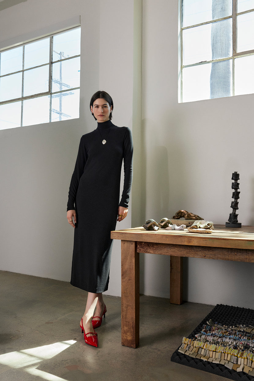 A person in the long black AGOURA DRESS by Velvet by Jenny Graham stands by a wooden table adorned with decorative items; bright red shoes provide a striking contrast to the minimalist room.