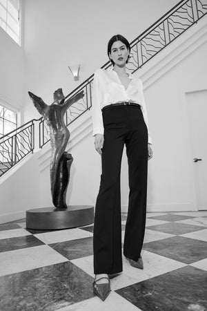 Inside near a staircase, someone is wearing the MARIPOSA PANT by Velvet by Jenny Graham teamed with a white top. An artistic sculpture can be seen in the background, enhancing the ambiance.