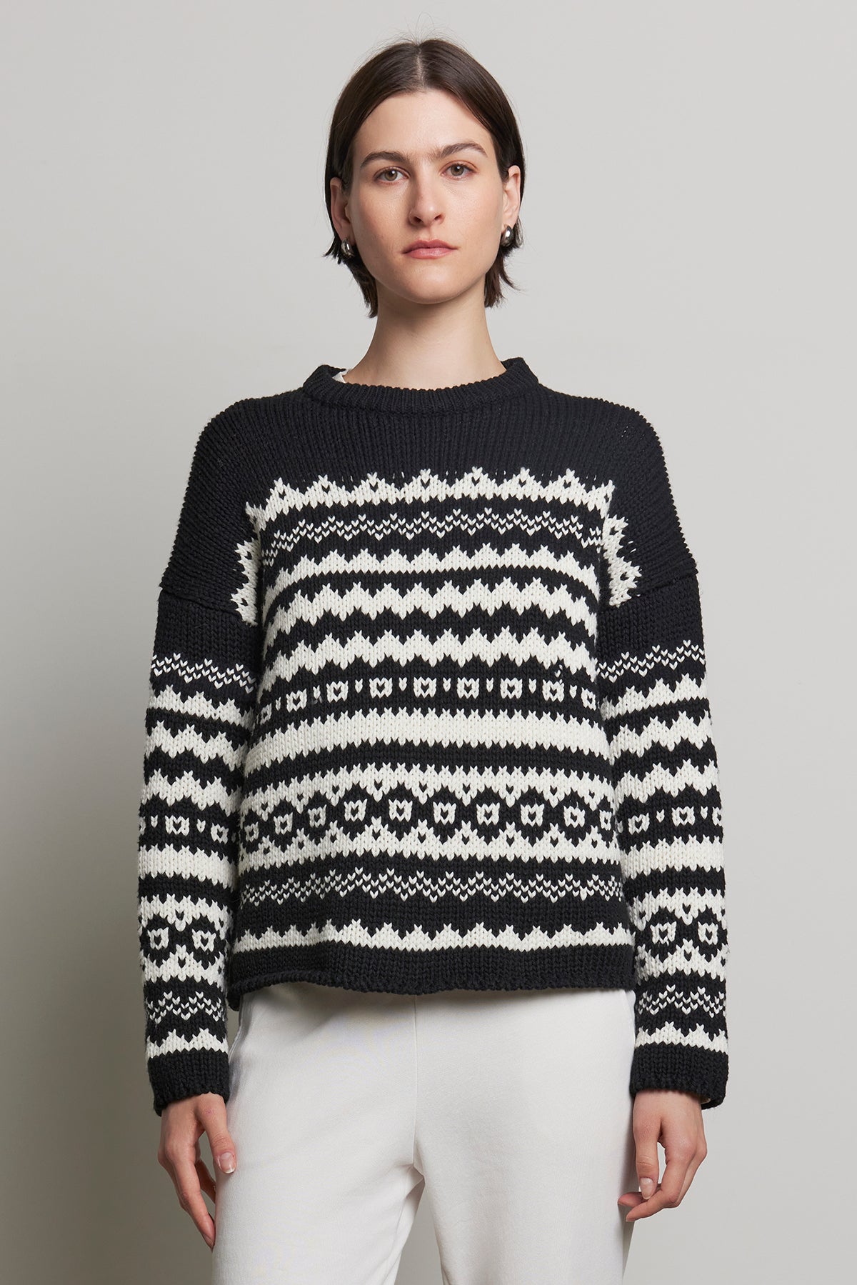   A person is seen wearing the MAYWOOD SWEATER by Velvet by Jenny Graham, featuring a relaxed fit and a black and white fair isle pattern, paired with white pants against a simple background. 