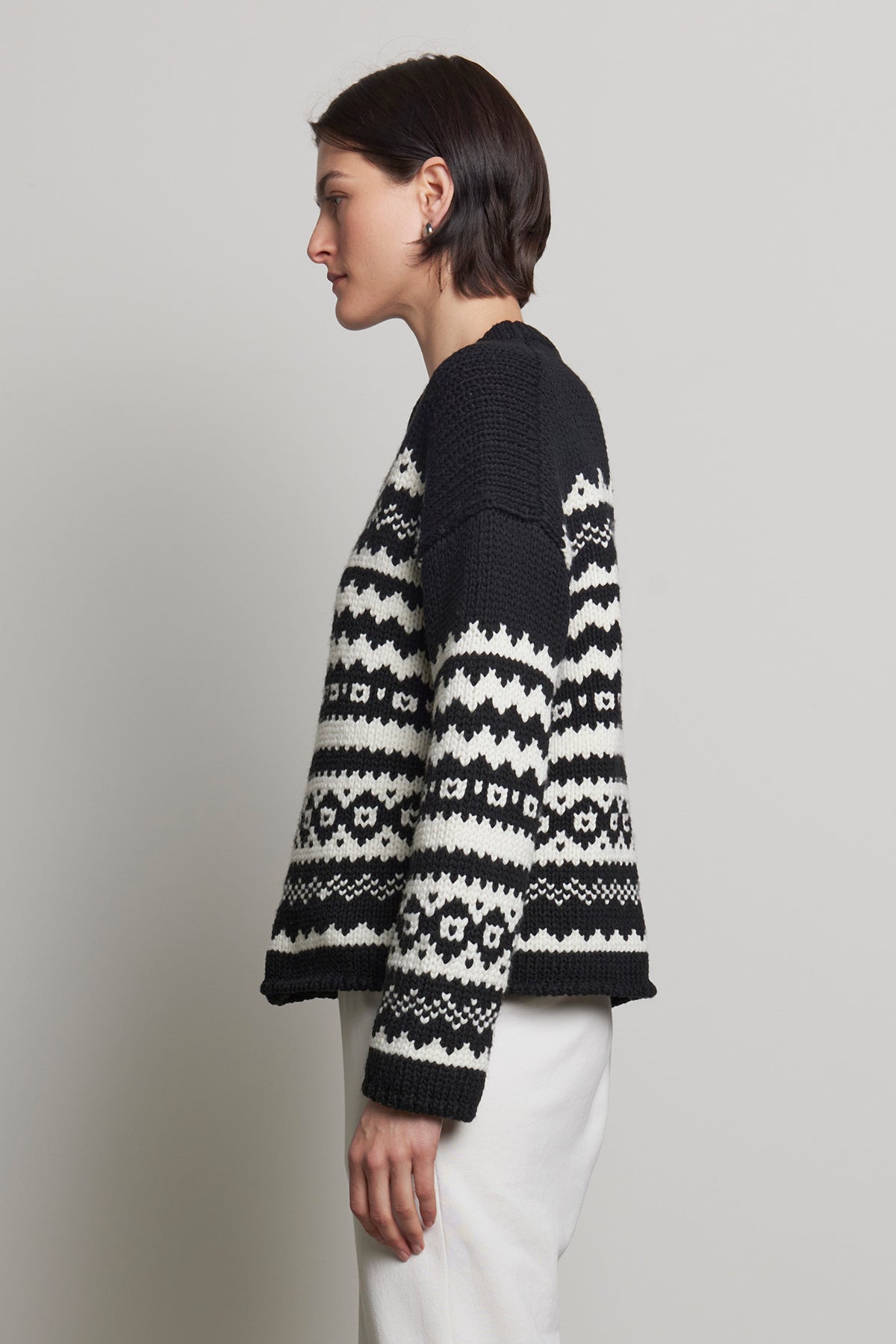   A person with short dark hair is wearing the MAYWOOD SWEATER by Velvet by Jenny Graham, featuring a black and white Fair Isle pattern crafted from a wool blend knit, paired with white pants, standing in profile against a plain background. 