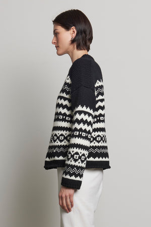 A person with short dark hair is wearing the MAYWOOD SWEATER by Velvet by Jenny Graham, featuring a black and white Fair Isle pattern crafted from a wool blend knit, paired with white pants, standing in profile against a plain background.