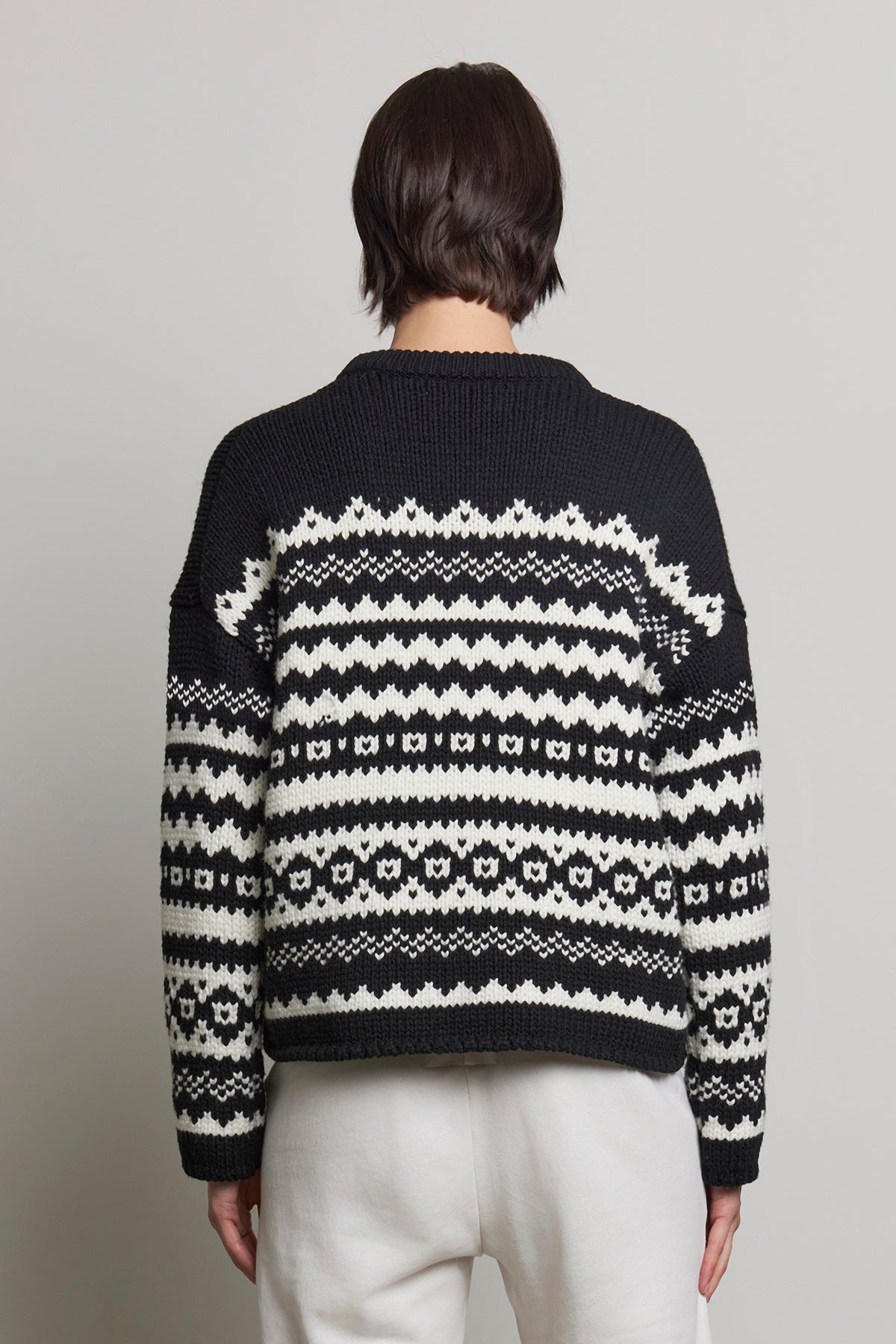   Rear view of a person wearing the MAYWOOD SWEATER by Velvet by Jenny Graham, featuring a black and white Fair Isle pattern, paired with white pants against a gray background. 