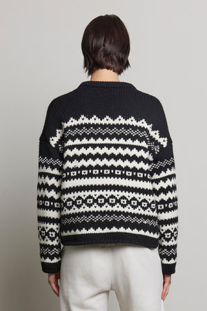 Rear view of a person wearing the MAYWOOD SWEATER by Velvet by Jenny Graham, featuring a black and white Fair Isle pattern, paired with white pants against a gray background.