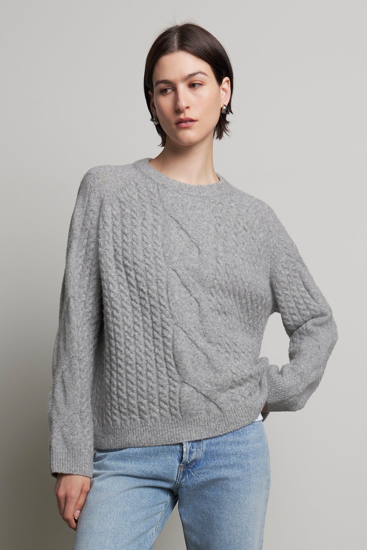   A person wearing the WILSHIRE SWEATER by Velvet by Jenny Graham, masterfully woven from a luxurious wool-yak blend in gray cable-knit, pairs it elegantly with blue jeans and stands against a plain background. 