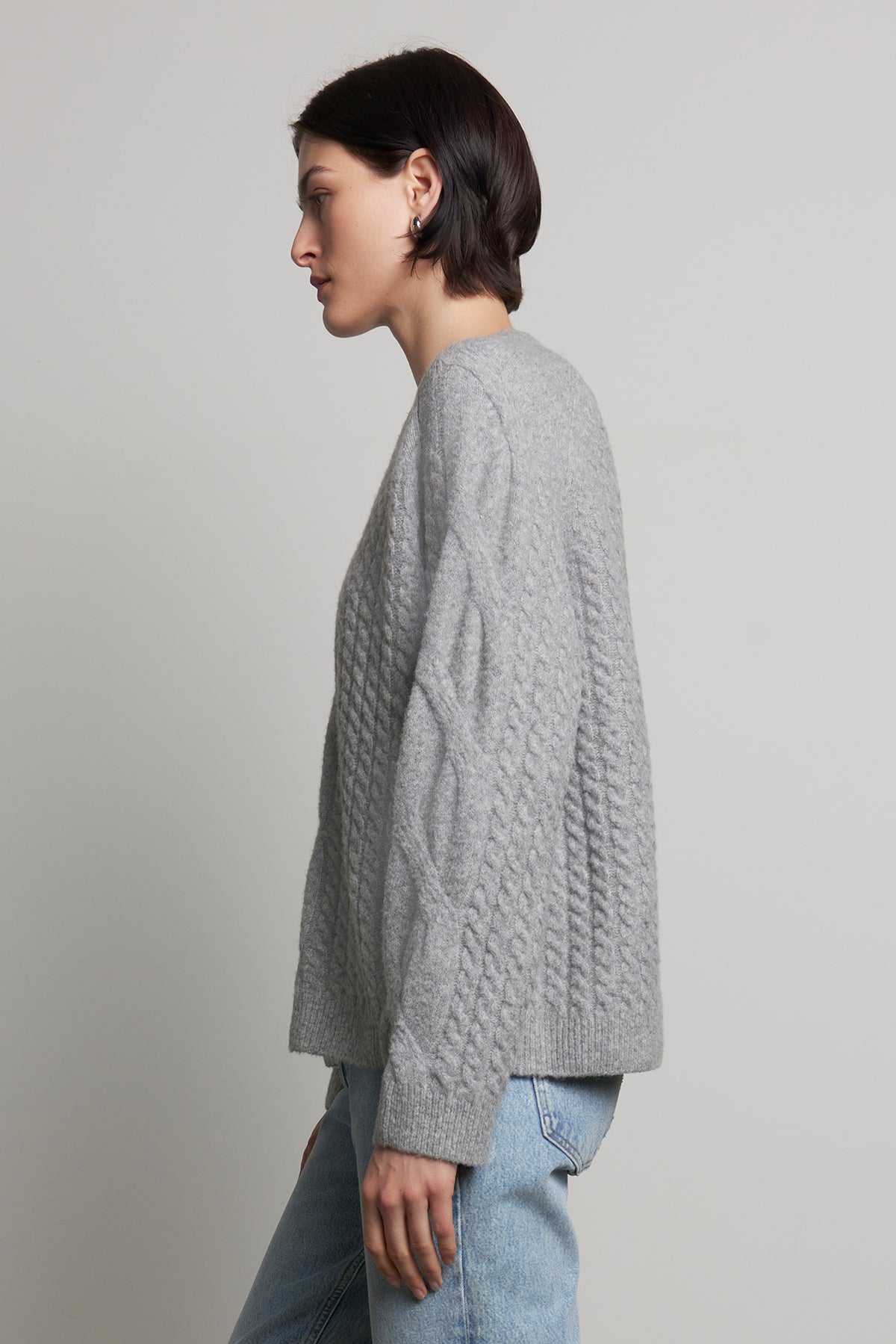   A person wearing the WILSHIRE SWEATER, a gray wool-yak blend cable-knit piece by Velvet by Jenny Graham, paired with jeans, stands against a plain background, facing left. 