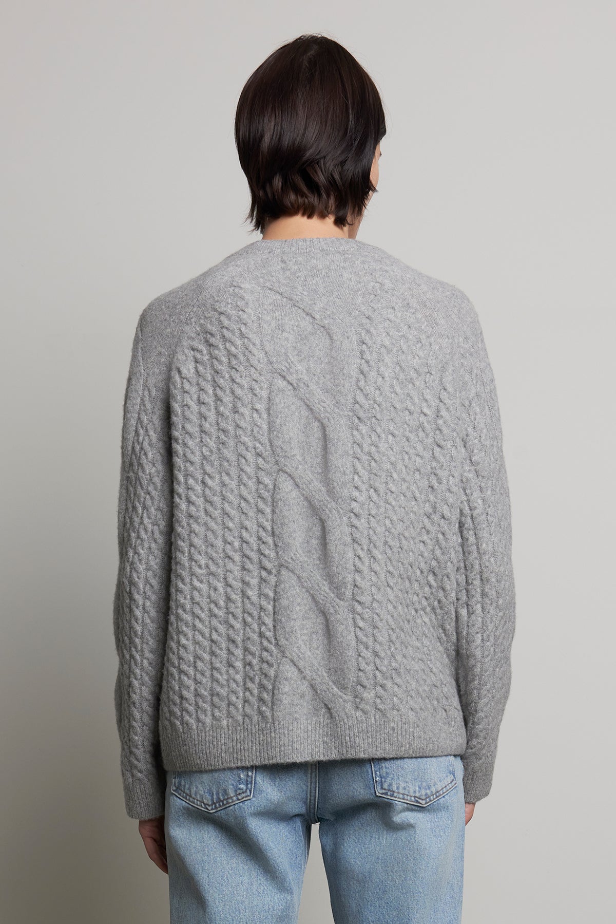 A person is photographed from the back against a neutral background wearing the WILSHIRE SWEATER, a gray wool-yak blend cable-knit piece by Velvet by Jenny Graham, paired with light blue jeans.-38318578401473