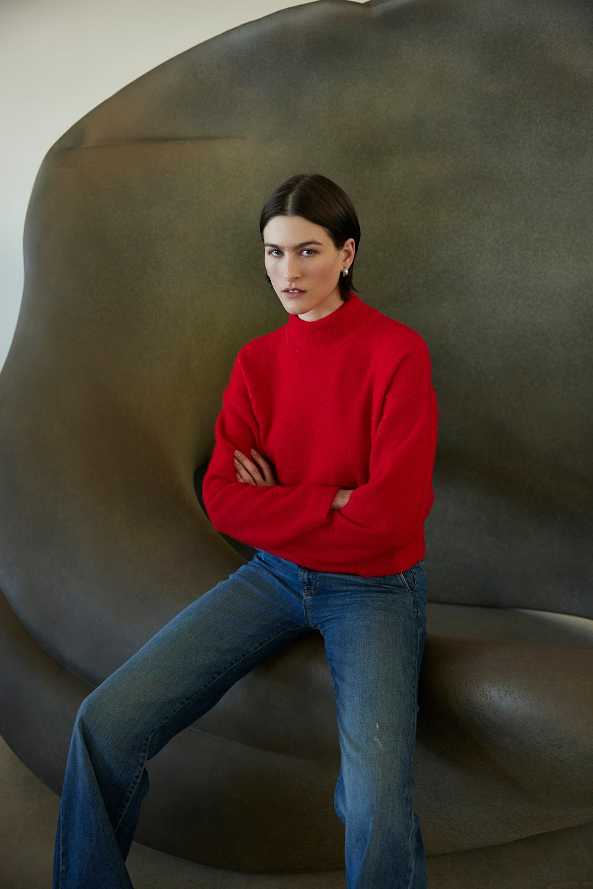 A person wearing a CYPRESS SWEATER by Velvet by Jenny Graham and jeans sits with arms crossed on a large, abstract gray sculpture.-38318581350593