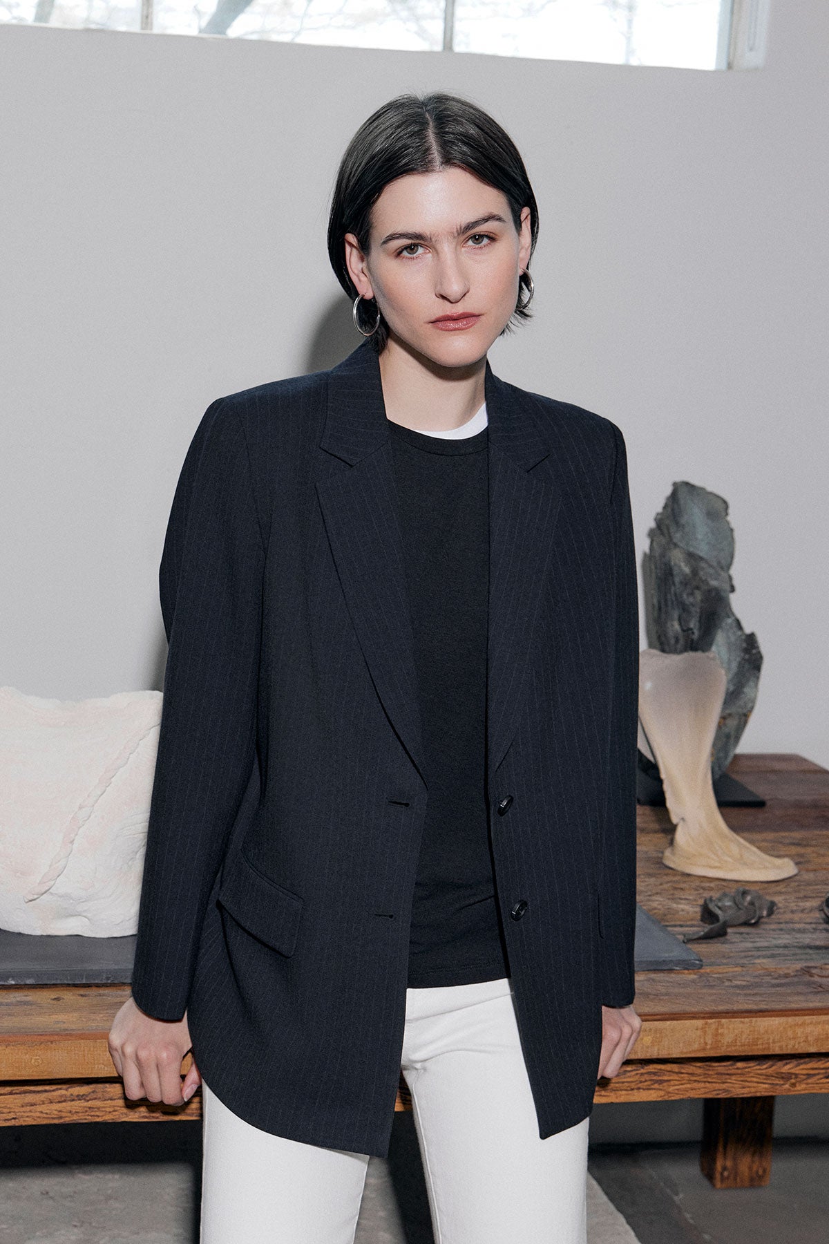 A person with short dark hair wears the FRESNO PINSTRIPE BLAZER by Velvet by Jenny Graham in an oversized fit, paired with a black shirt and white pants, standing indoors near a wooden table with decorative objects.-39274356539585