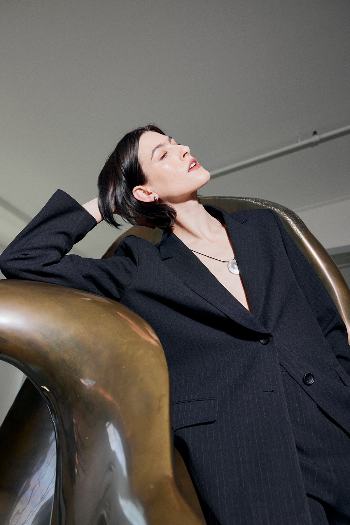   Dressed in the TAHOE PINSTRIPE PANT by Velvet by Jenny Graham, a person leans on a brass sculpture, gazing upward. Their short dark hair and pendant necklace complement the refined look of their attire. 