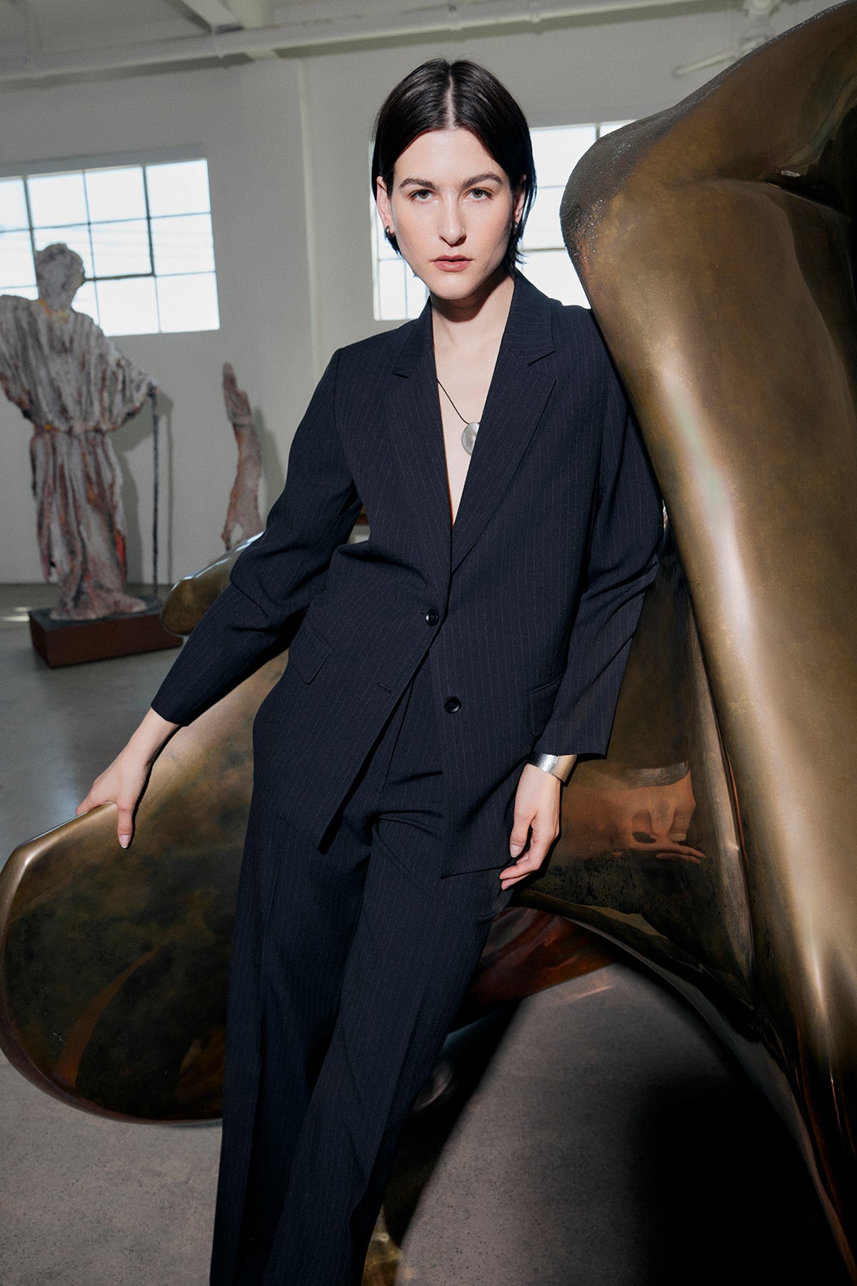   A person wearing the FRESNO PINSTRIPE BLAZER from Velvet by Jenny Graham stands in front of a large abstract sculpture in a well-lit room with industrial windows. 