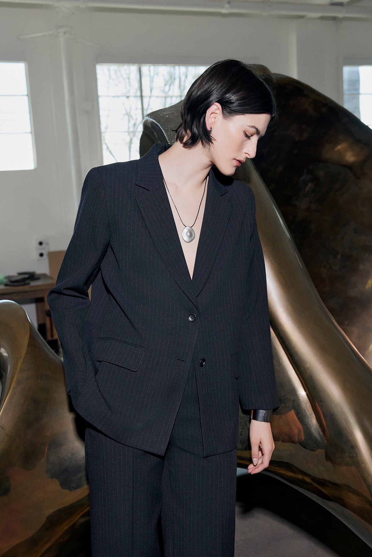   A person wearing a Velvet by Jenny Graham FRESNO PINSTRIPE BLAZER with a notched collar and a necklace stands indoors near a large abstract sculpture. 