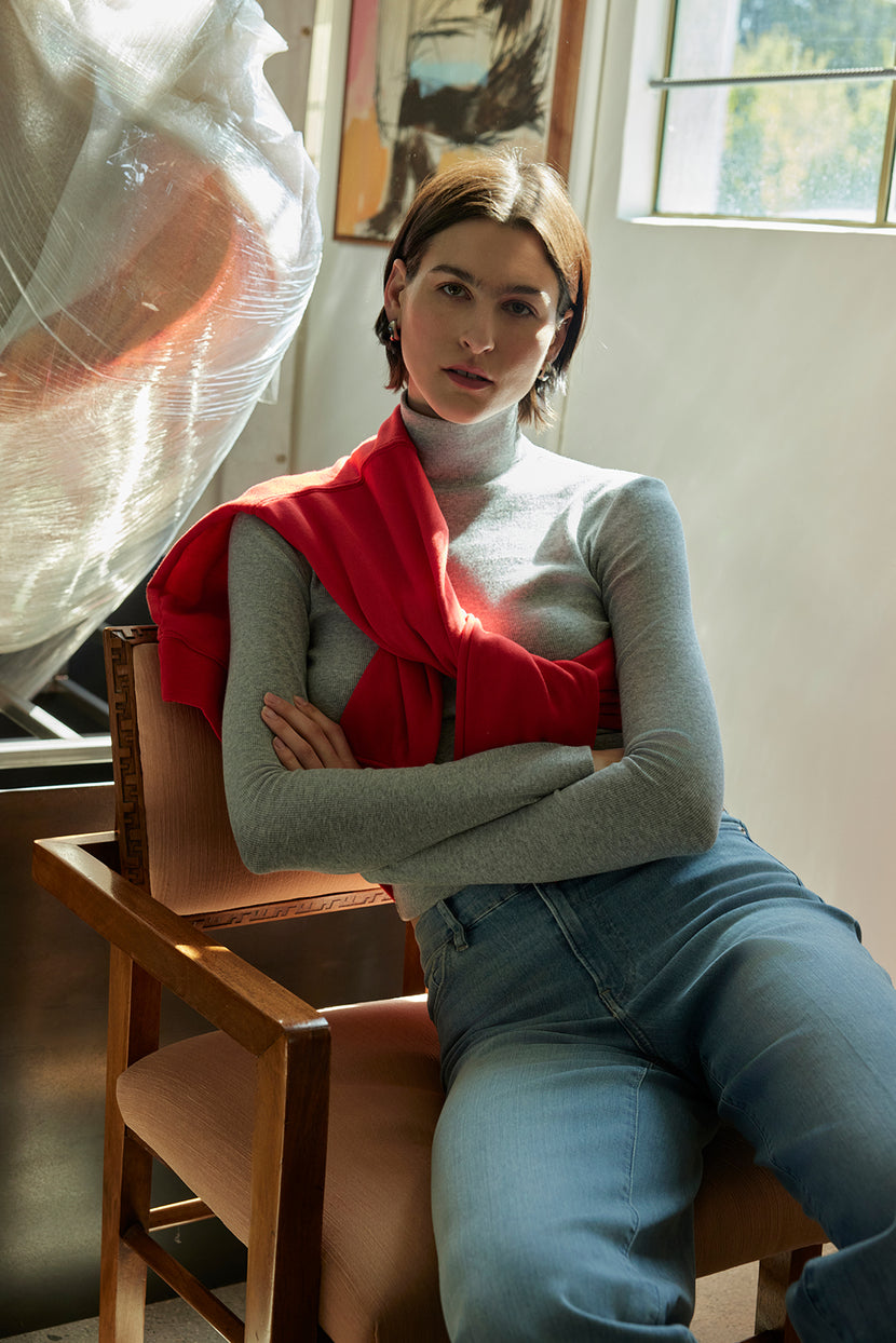 A person sits on a chair in a sunlit room, wearing a gray top and jeans. The YNEZ SWEATSHIRT by Velvet by Jenny Graham, made from organic cotton in vibrant red, is draped over their shoulders, seamlessly blending style with sustainable fashion. Art adds a creative touch to the background.