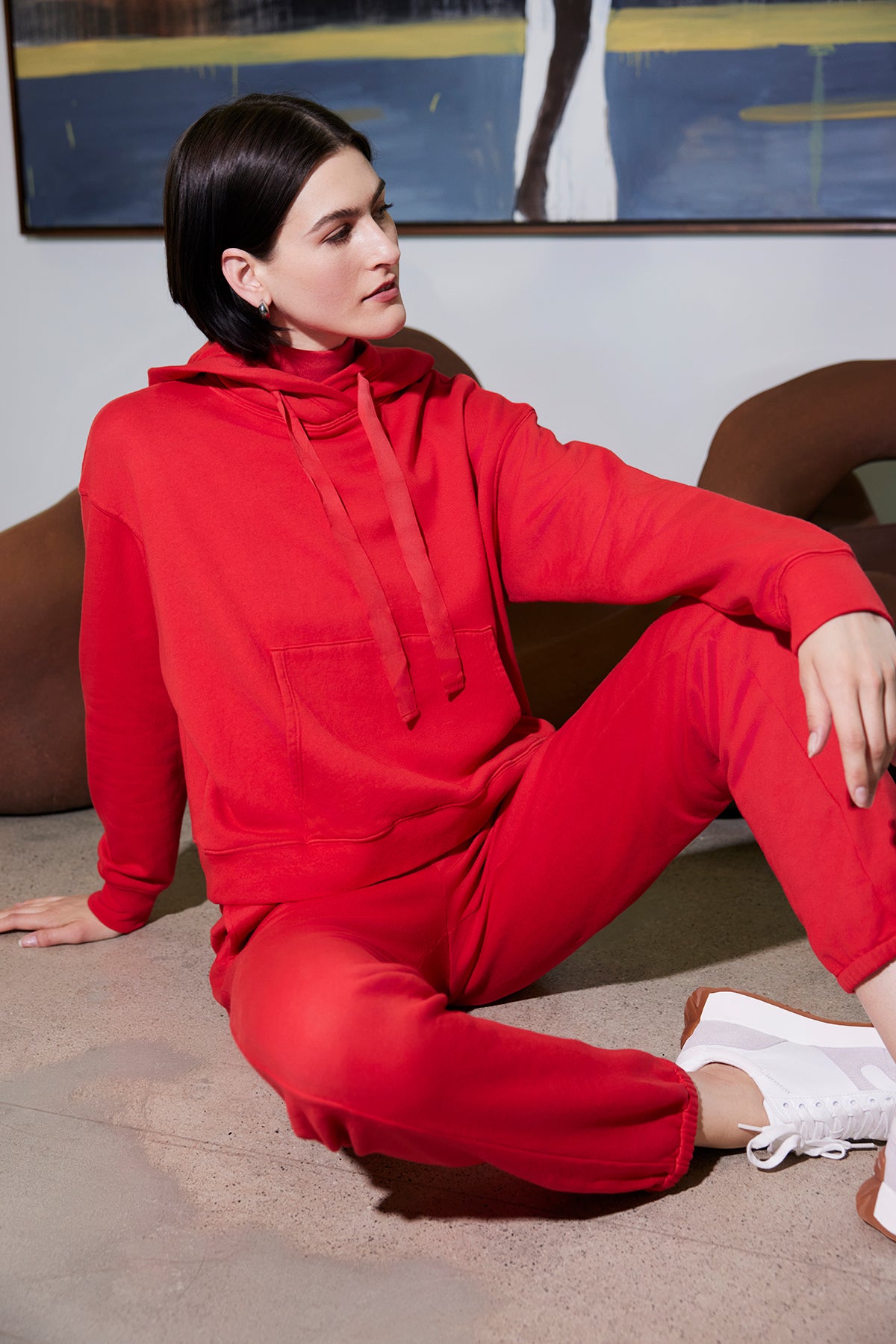   A person wearing a vibrant red hoodie and the ZUMA SWEATPANT by Velvet by Jenny Graham sits on the floor, embodying sustainable fashion. 