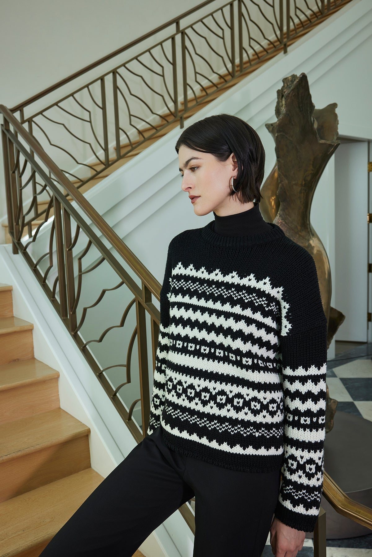   A person wearing the Velvet by Jenny Graham MAYWOOD SWEATER, featuring a relaxed fit and chunky wool blend knit with a fair isle pattern, stands by a staircase with a wooden railing. 