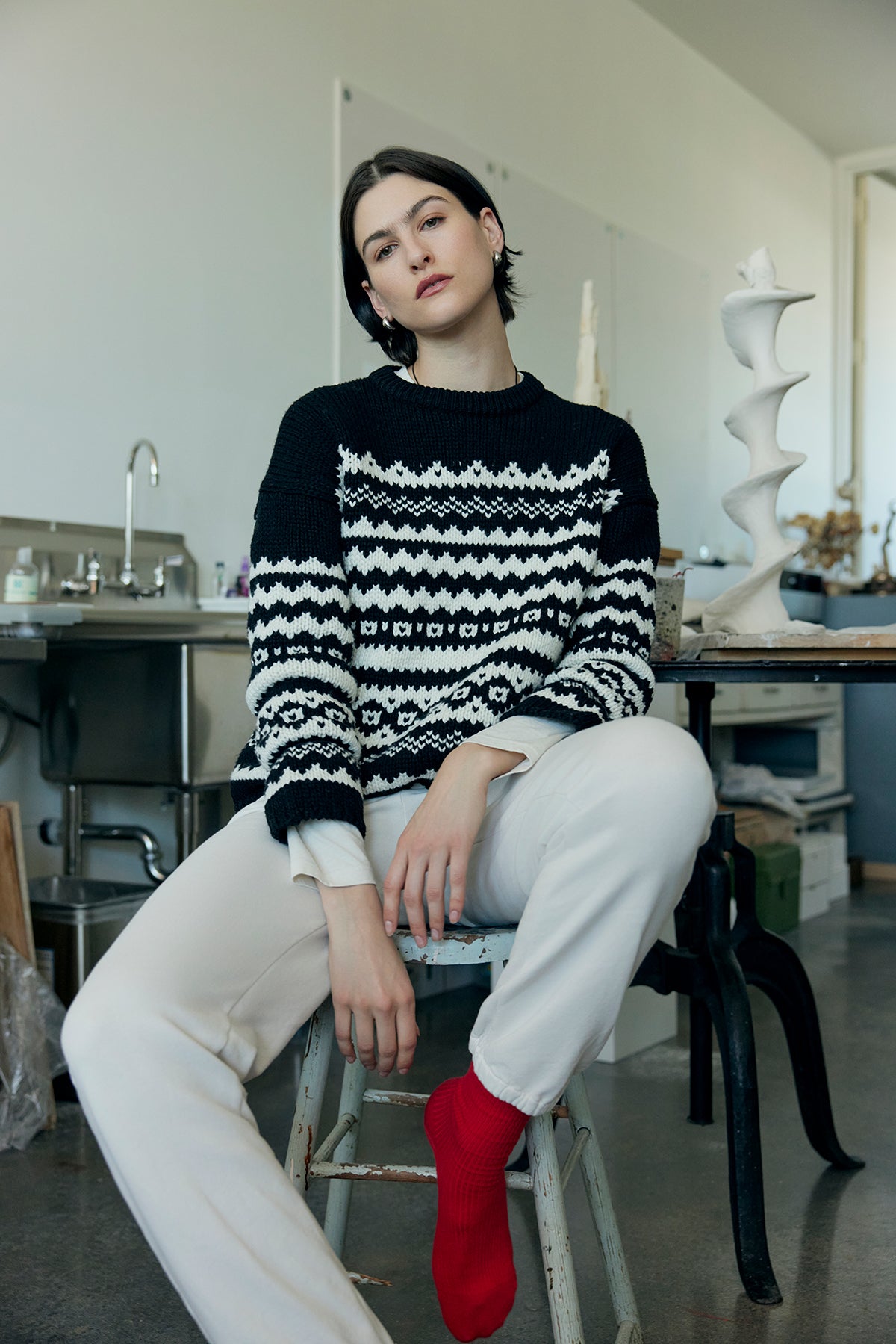 In a room adorned with shelves and sculptures, a person sits on a stool wearing the MAYWOOD SWEATER by Velvet by Jenny Graham, which features an intricate fair isle pattern. This relaxed-fit sweater is paired with white pants and vibrant red socks.-38318585053377