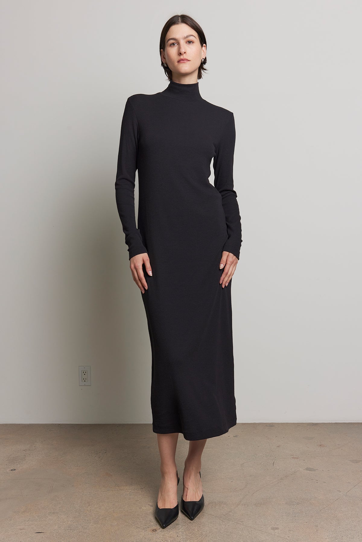 A person wearing the AGOURA DRESS by Velvet by Jenny Graham, styled with black pointed shoes, stands against a plain background.-38318580465857