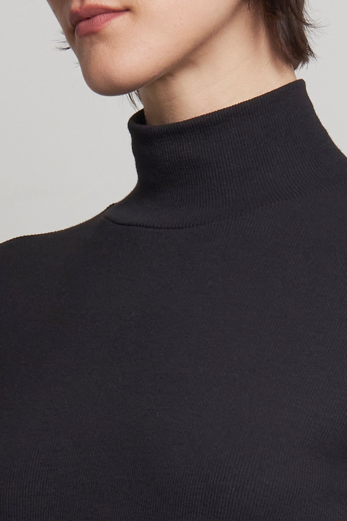   Close-up of a person wearing the AGOURA DRESS by Velvet by Jenny Graham, featuring a fitted silhouette and ribbed cotton black turtleneck design, highlighting their chin and neck. 