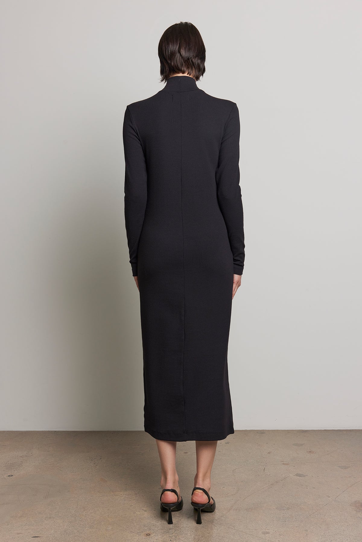 With their short hair turned away from the camera, the individual is wearing the AGOURA DRESS, a sleek long-sleeved black midi dress by Velvet by Jenny Graham, paired with black heels.-38318580531393