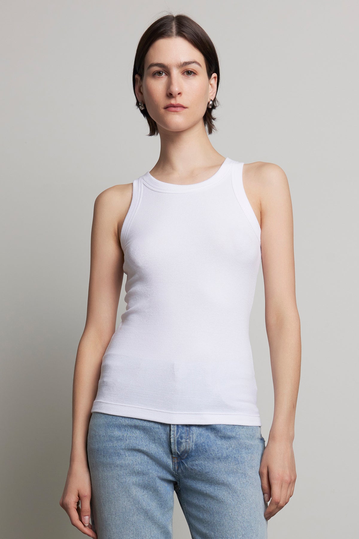   A person with short hair wearing the CRUZ TANK TOP by Velvet by Jenny Graham, crafted from ribbed cotton-modal, and blue jeans stands against a plain background, embodying the essence of a capsule wardrobe. 