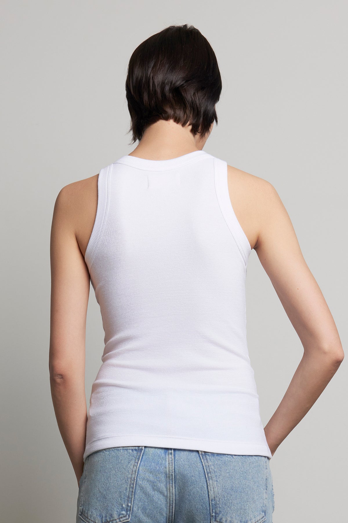   Rear view of a person with short hair wearing the CRUZ TANK TOP by Velvet by Jenny Graham, paired with blue jeans, highlighting a capsule wardrobe essential against a plain background. 