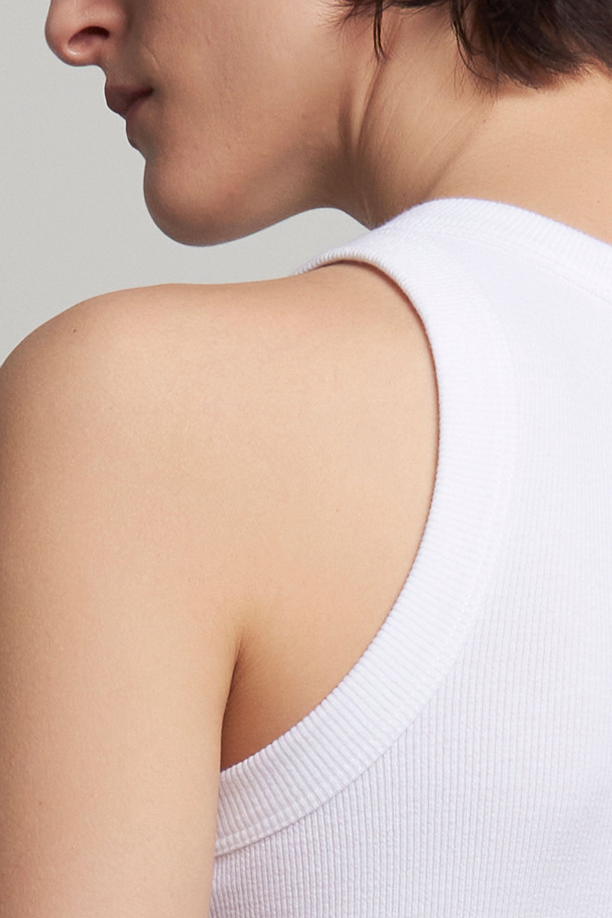 Close-up of a person wearing the Velvet by Jenny Graham CRUZ TANK TOP in a white cotton-modal ribbed blend. The shoulder and neck are shown, with the face partially visible and turned away—an ideal piece for any capsule wardrobe.-40101680939201