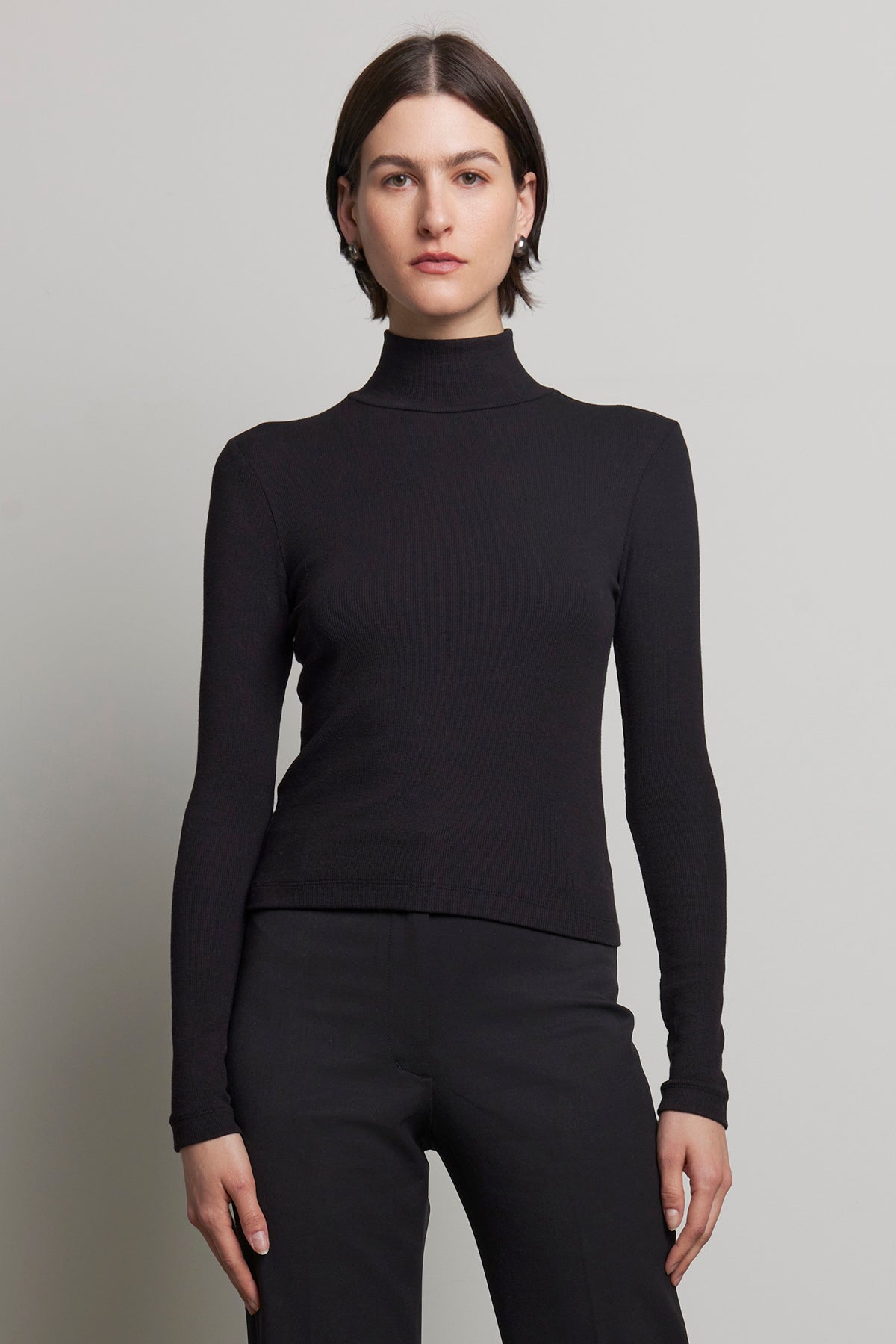   A person stands against a neutral background, dressed in the sleek HELMS TEE by Velvet by Jenny Graham, featuring a black long-sleeve turtleneck with a subtle ribbed texture and matching pants. 